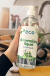 A bottle of eco dishwashing soap held in front of a sink full of dishes, for an eco-friendly and sustainable kitchen.